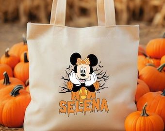 May include: A white canvas tote bag with a Mickey Mouse character dressed as a pumpkin. The bag has the name "SELENA" written in orange dripping letters. The bag is surrounded by orange pumpkins.