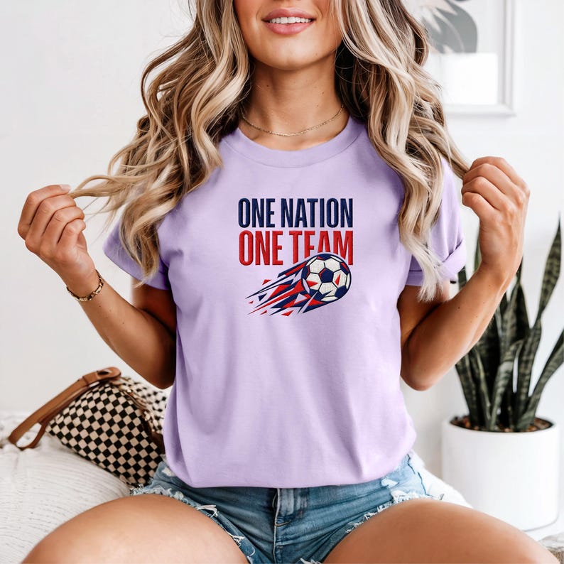 May include: A light purple t-shirt with the text "ONE NATION ONE TEAM" in navy blue and red. A soccer ball graphic with red and blue streaks is below the text. The shirt is made of a soft material.