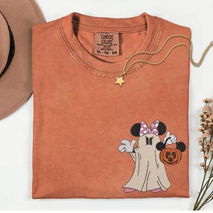 Pu&ograve; includere: T-shirt Comfort Colors color ruggine con un design a fantasma di Minnie Mouse che tiene in mano una lanterna a forma di zucca. La maglietta ha un girocollo e maniche corte. Una collana con pendente a stella dorata &egrave; drappeggiata sulla maglietta. Un cappello marrone &egrave; nell'angolo in alto a sinistra.