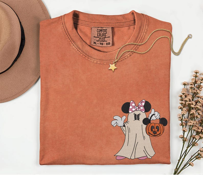Pu&ograve; includere: T-shirt Comfort Colors color ruggine con un design a fantasma di Minnie Mouse che tiene in mano una lanterna a forma di zucca. La maglietta ha un girocollo e maniche corte. Una collana con pendente a stella dorata &egrave; drappeggiata sulla maglietta. Un cappello marrone &egrave; nell'angolo in alto a sinistra.