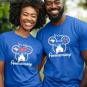 May include: Two people wearing matching blue t-shirts. The shirts feature a white graphic of Mickey Mouse ears with hearts, a bow, and a castle, along with the text "It's our 1st Anniversary."
