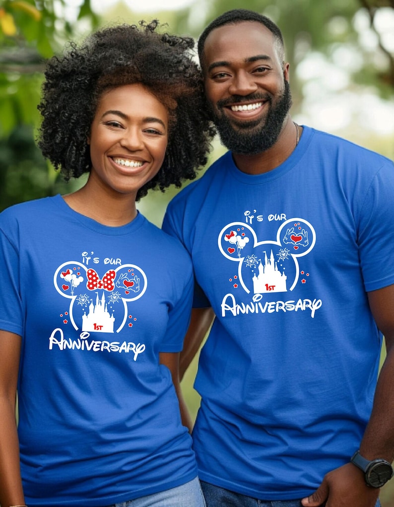 May include: Two people wearing matching blue t-shirts. The shirts feature a white graphic of Mickey Mouse ears with hearts, a bow, and a castle, along with the text "It's our 1st Anniversary."
