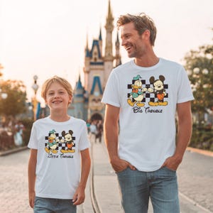 May include: White t-shirts with a cartoon graphic of Donald Duck and Mickey Mouse on a checkered background. The shirt on the left says "Little Trouble" and the shirt on the right says "Big Trouble".