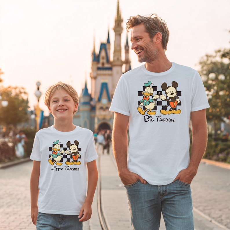 May include: White t-shirts with a cartoon graphic of Donald Duck and Mickey Mouse on a checkered background. The shirt on the left says "Little Trouble" and the shirt on the right says "Big Trouble".