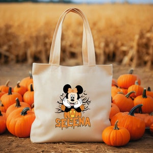 May include: A white canvas tote bag with a Halloween-themed design featuring Mickey Mouse dressed as a pumpkin. The bag has the name "SELENA" printed in orange dripping letters below the image.