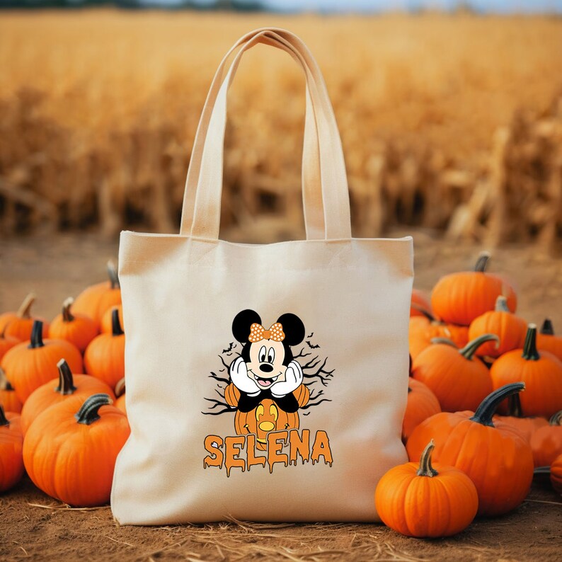 May include: A white canvas tote bag with a Halloween-themed design featuring Mickey Mouse dressed as a pumpkin. The bag has the name "SELENA" printed in orange dripping letters below the image.