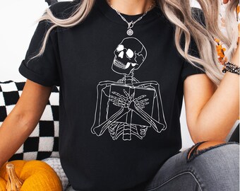 May include: A black short-sleeved t-shirt featuring a white line art graphic of a skeleton holding a book against its chest. The skeleton is depicted from the ribcage up, with its hands clasped over the cover of the book, and its head tilted.