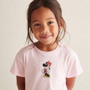 May include: A light pink t-shirt featuring a Minnie Mouse graphic. The graphic shows Minnie Mouse in a red and white polka dot dress and bow, with yellow shoes. The shirt is made of a soft material.
