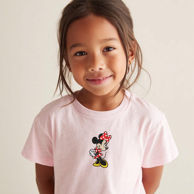May include: A light pink t-shirt featuring a Minnie Mouse graphic. The graphic shows Minnie Mouse in a red and white polka dot dress and bow, with yellow shoes. The shirt is made of a soft material.