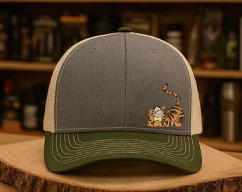 May include: A gray and khaki trucker hat with a green brim. The hat features an embroidered image of Tigger from Winnie the Pooh. The hat is sitting on a wooden surface.