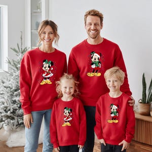 K&ouml;nnte beinhalten: Rote Sweatshirts mit einem Weihnachtsdesign von Minnie Mouse und Mickey Mouse. Die Sweatshirts werden von einer vierk&ouml;pfigen Familie getragen. Das Minnie Mouse-Design hat eine gr&uuml;ne Schleife und gelbe Schuhe. Das Mickey Mouse-Design hat einen Weihnachtsmannhut und einen gr&uuml;nen Schal.