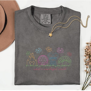 May include: A gray Comfort Colors t-shirt with a colorful embroidered design of a castle, sphere, and tree, with fireworks and the text "what a wonderful world." A gold star pendant necklace rests on the shirt.