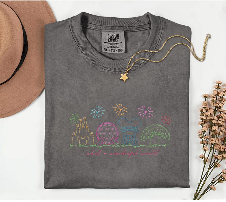 May include: A gray Comfort Colors t-shirt with a colorful embroidered design of a castle, sphere, and tree, with fireworks and the text "what a wonderful world." A gold star pendant necklace rests on the shirt.