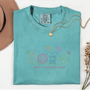 May include: A turquoise Comfort Colors XL t-shirt with a gold star necklace. The shirt features a colorful line drawing of a theme park with fireworks and the text "...what a wonderful world."