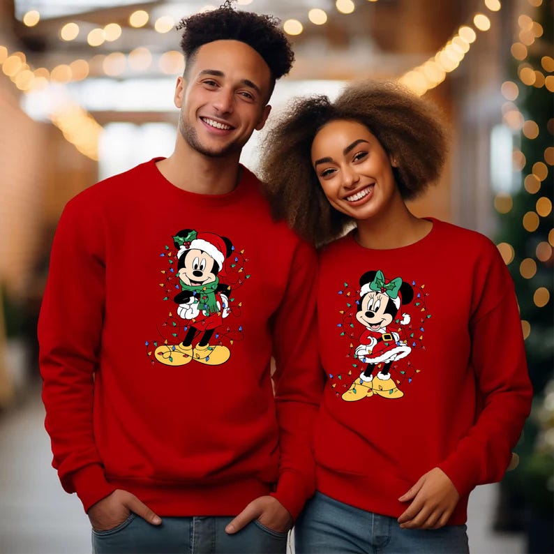 K&ouml;nnte beinhalten: Zwei rote Sweatshirts mit Mickey und Minnie Mouse in Weihnachtskleidung. Mickey tr&auml;gt einen Weihnachtsmannhut, einen gr&uuml;nen Schal und gelbe Schuhe. Minnie tr&auml;gt eine gr&uuml;ne Schleife, ein rotes Kleid und gelbe Schuhe, umgeben von Lichterketten.