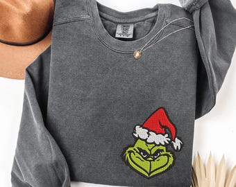 May include: A charcoal gray sweatshirt featuring an embroidered Grinch face wearing a Santa hat. The sweatshirt has a crew neck and long sleeves. A gold necklace with a pendant is draped on the shirt.