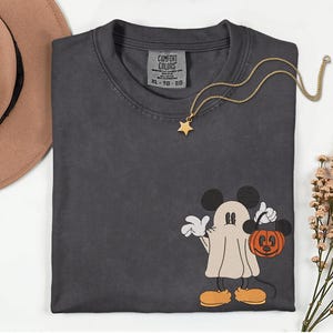 Pu&ograve; includere: T-shirt Comfort Colors grigio scuro con un design di Mickey Mouse a tema Halloween. Mickey &egrave; vestito da fantasma e tiene in mano una zucca. Una collana con pendente a stella dorata &egrave; drappeggiata sulla maglietta. Un cappello marrone e dei fiori sono sullo sfondo.