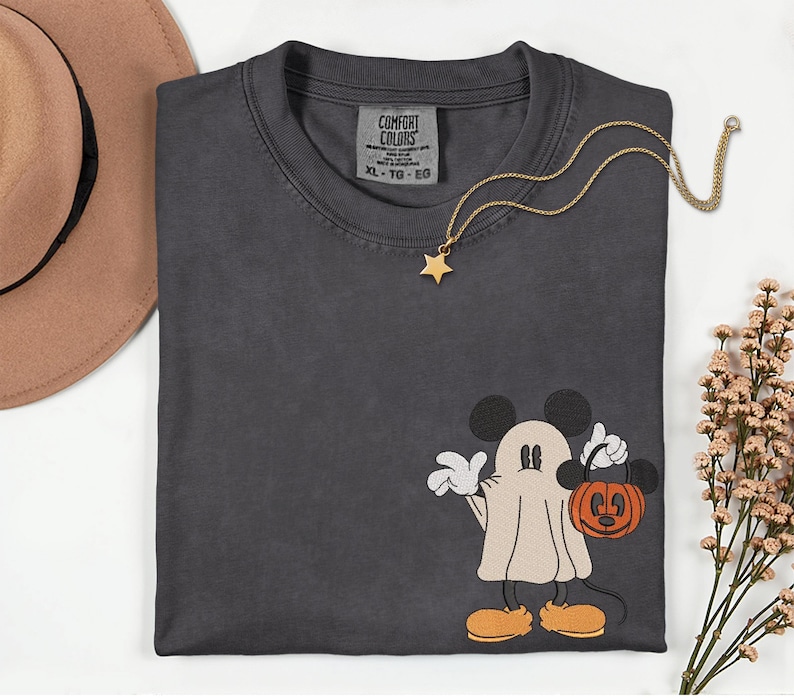 Pu&ograve; includere: T-shirt Comfort Colors grigio scuro con un design di Mickey Mouse a tema Halloween. Mickey &egrave; vestito da fantasma e tiene in mano una zucca. Una collana con pendente a stella dorata &egrave; drappeggiata sulla maglietta. Un cappello marrone e dei fiori sono sullo sfondo.