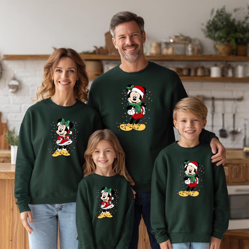 K&ouml;nnte beinhalten: Vier Personen tragen passende dunkelgr&uuml;ne Sweatshirts. Jedes Sweatshirt zeigt eine Cartoon-Figur von Minnie Mouse mit Weihnachtsm&uuml;tze und Weihnachtsoutfit, umgeben von bunten Lichtern. Die Familie l&auml;chelt.