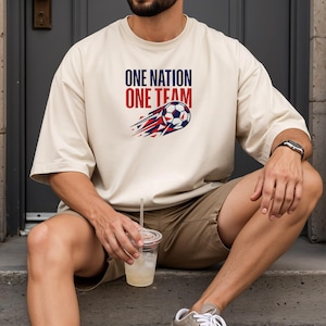 May include: Cream-colored t-shirt with the text "ONE NATION ONE TEAM" above a soccer ball graphic with red, white, and blue accents. The shirt is worn by a person sitting on steps.