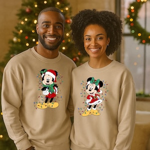 K&ouml;nnte beinhalten: Zwei Personen tragen passende beige Sweatshirts mit weihnachtlichen Mickey- und Minnie-Mouse-Designs. Mickey tr&auml;gt einen Weihnachtsmannhut und Schal, Minnie ein rotes Kleid und eine Schleife, beide mit Lichterketten.