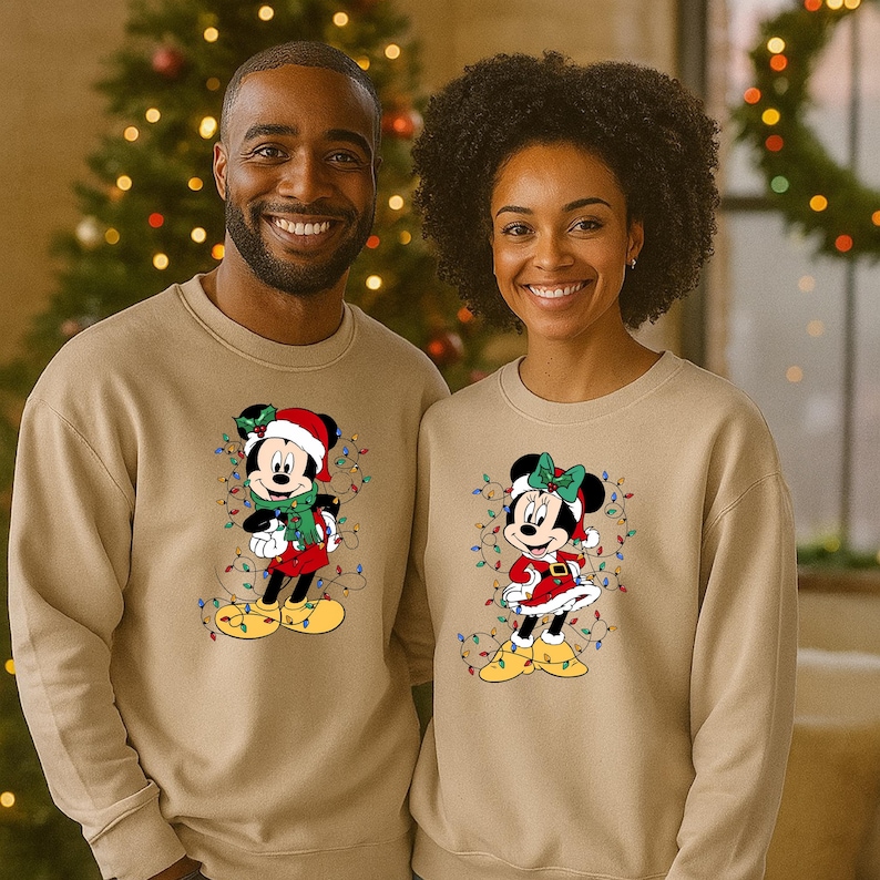 K&ouml;nnte beinhalten: Zwei Personen tragen passende beige Sweatshirts mit weihnachtlichen Mickey- und Minnie-Mouse-Designs. Mickey tr&auml;gt einen Weihnachtsmannhut und Schal, Minnie ein rotes Kleid und eine Schleife, beide mit Lichterketten.