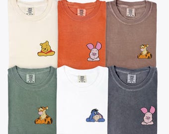May include: Six different colored t-shirts, each with a small embroidered cartoon character. Colors include ivory, yam, espresso, moss, white, and pepper. The characters are Winnie the Pooh, Piglet, Tigger, and Eeyore.