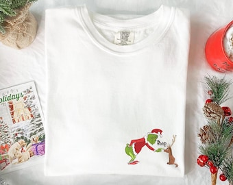May include: White t-shirt with a crew neck featuring a cartoon Grinch character in a Santa hat, touching the nose of a reindeer. The word "Boop" is above the reindeer. A holiday card and Christmas decorations are in the background.