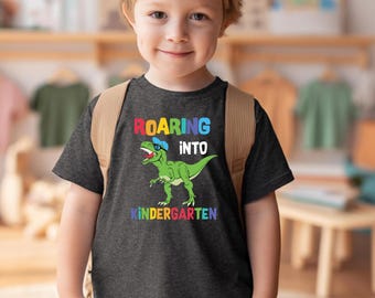 May include: A dark gray short-sleeved t-shirt featuring a green dinosaur wearing blue sunglasses and a blue cap. The text "ROARING INTO KINDERGARTEN" is printed in colorful letters above and below the dinosaur graphic on the front of the shirt.