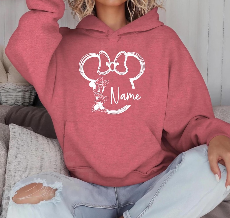 K&ouml;nnte beinhalten: Ein roter Kapuzenpullover mit einer wei&szlig;en Umrandung von Minnie Mauss Kopf und dem Wort "Name" auf der Vorderseite.