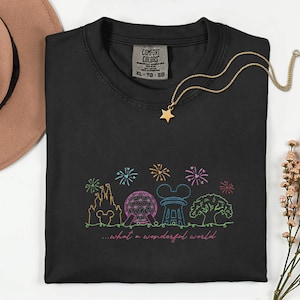 May include: Black Comfort Colors t-shirt with a colorful embroidered design of a castle, Spaceship Earth, Mickey Mouse ears, and a tree, with fireworks and the text "...what a wonderful world." A gold star necklace is draped on the shirt.