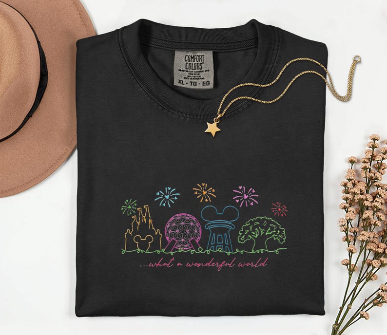 May include: Black Comfort Colors t-shirt with a colorful embroidered design of a castle, Spaceship Earth, Mickey Mouse ears, and a tree, with fireworks and the text "...what a wonderful world." A gold star necklace is draped on the shirt.