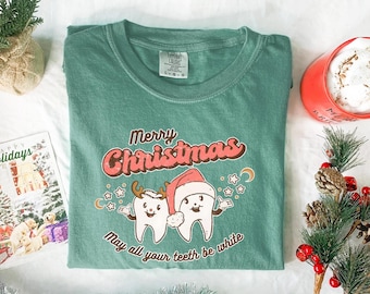 May include: A green t-shirt with a Christmas-themed graphic. The graphic features two cartoon teeth wearing a Santa hat and antlers, with the text "Merry Christmas" and "May all your teeth be white."