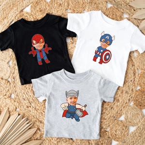 K&ouml;nnte beinhalten: Drei T-Shirts mit verschiedenen Superhelden-Designs. Das erste Shirt ist schwarz mit einem roten und blauen Spiderman-Design. Das zweite Shirt ist wei&szlig; mit einem blauen und roten Captain America-Design. Das dritte Shirt ist grau mit einem roten und gelben Thor-Design.
