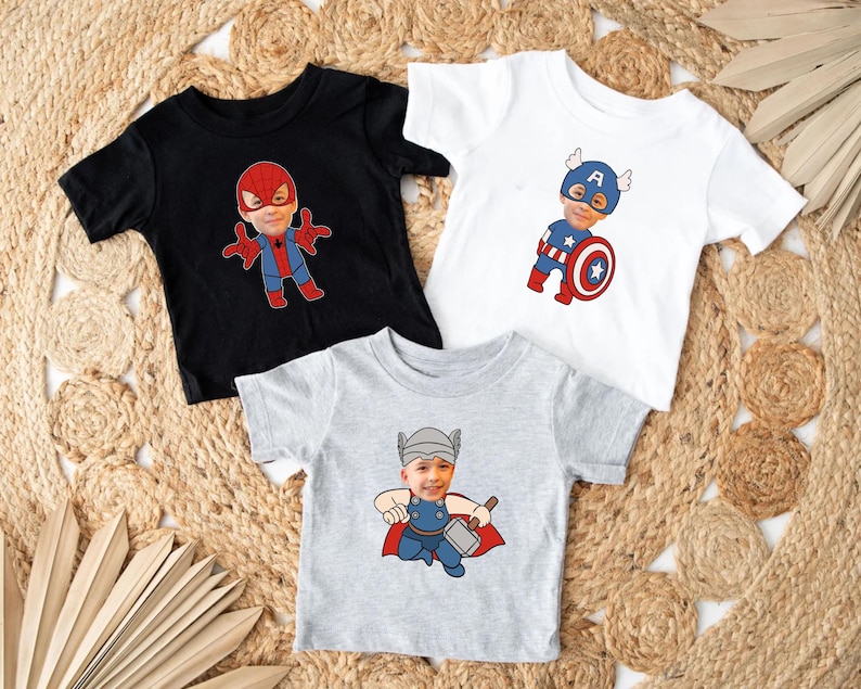 K&ouml;nnte beinhalten: Drei T-Shirts mit verschiedenen Superhelden-Designs. Das erste Shirt ist schwarz mit einem roten und blauen Spiderman-Design. Das zweite Shirt ist wei&szlig; mit einem blauen und roten Captain America-Design. Das dritte Shirt ist grau mit einem roten und gelben Thor-Design.