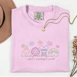 May include: A light pink Comfort Colors t-shirt with a colorful embroidered design of a castle, sphere, and other icons. The text "...what a wonderful world" is below the design. A gold star pendant necklace is on the shirt.