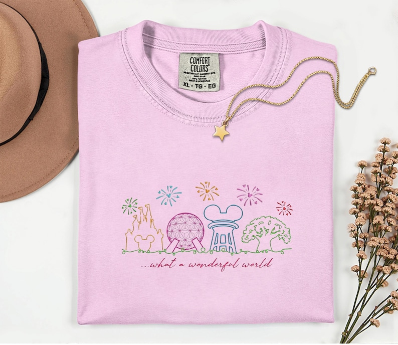 May include: A light pink Comfort Colors t-shirt with a colorful embroidered design of a castle, sphere, and other icons. The text "...what a wonderful world" is below the design. A gold star pendant necklace is on the shirt.