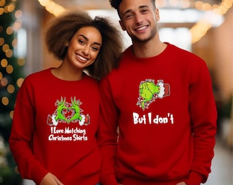 May include: A couple wearing matching red Christmas sweaters with a Grinch-themed design. The woman's sweater says "I love matching Christmas shirts" and the man's sweater says "But I don't".