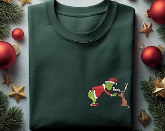 May include: Dark green sweatshirt featuring a cartoon Grinch character in a Santa hat, touching a reindeer's nose. The word "Boop" is next to the reindeer. Christmas-themed apparel.
