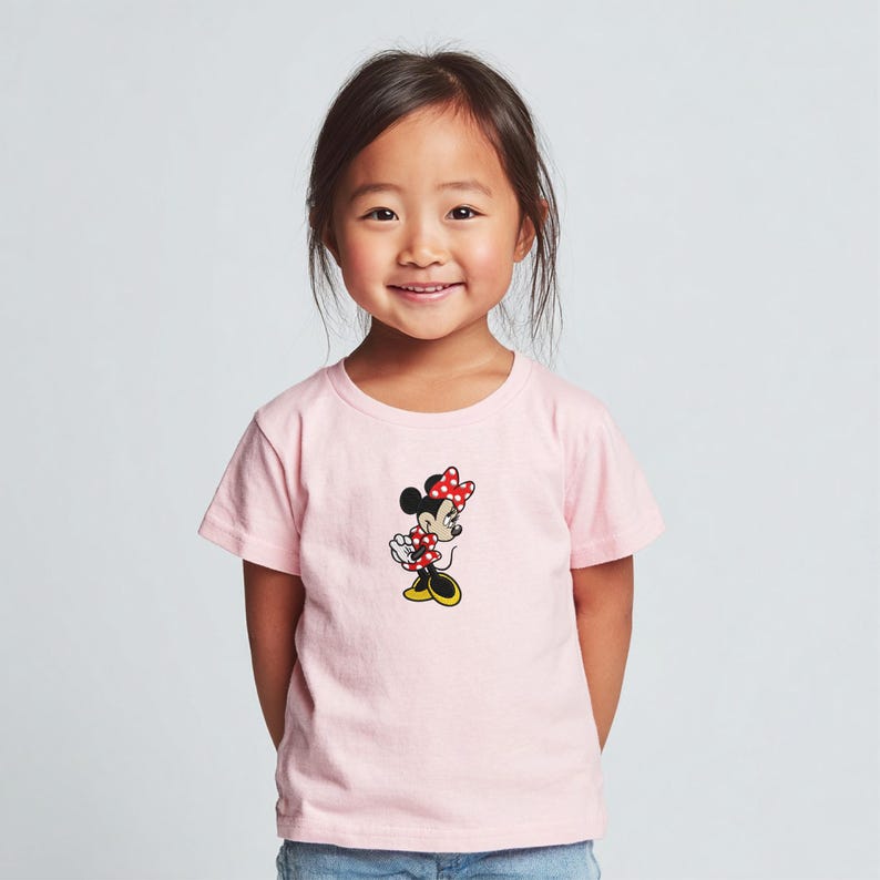 May include: A light pink t-shirt featuring a Minnie Mouse graphic. The cartoon character is depicted with a red and white polka dot bow, dress, and shoes. The shirt is short-sleeved and appears to be made of cotton.