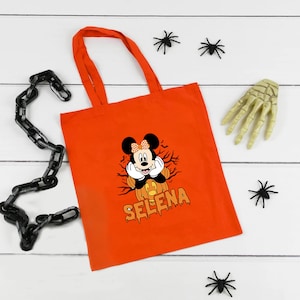 May include: Orange tote bag with a black and white image of Mickey Mouse dressed as a pumpkin. The text "Selena" is printed in black below the image.