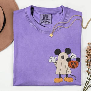 Pu&ograve; includere: Una maglietta Comfort Colors color lavanda con un design a fantasma di Topolino che tiene in mano una zucca. La maglietta ha uno scollo tondo e un'etichetta sul collo. Una collana con pendente a stella dorata &egrave; drappeggiata sulla maglietta.