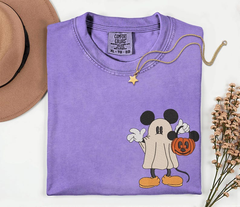 Pu&ograve; includere: Una maglietta Comfort Colors color lavanda con un design a fantasma di Topolino che tiene in mano una zucca. La maglietta ha uno scollo tondo e un'etichetta sul collo. Una collana con pendente a stella dorata &egrave; drappeggiata sulla maglietta.