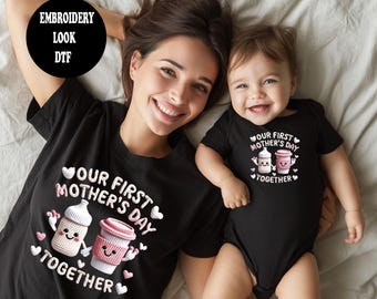 May include: A mother and baby wearing matching black t-shirts and onesie. The shirts and onesie have the text "OUR FIRST MOTHER'S DAY TOGETHER" with illustrations of a baby bottle and a coffee cup. The image is taken from above.