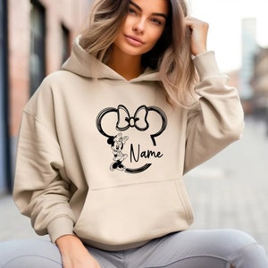 K&ouml;nnte beinhalten: Ein beiger Hoodie mit einem schwarz-wei&szlig;en Motiv von Minnie Mauss Kopf mit Schleife und dem Wort "Name" darunter.