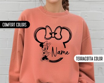 May include: A terracotta colored sweatshirt with a black and white graphic of Minnie Mouse's head and the word "Name" underneath. The sweatshirt is a comfortable fit and has a crew neck.
