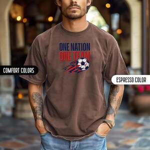 May include: Espresso-colored t-shirt with the text "ONE NATION ONE TEAM" above a soccer ball graphic with red and blue streaks. The shirt has short sleeves and a crew neck. The words "COMFORT COLORS" are on the left side of the image.