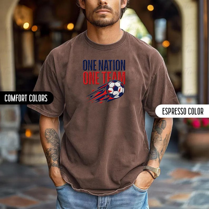 May include: Espresso-colored t-shirt with the text "ONE NATION ONE TEAM" above a soccer ball graphic with red and blue streaks. The shirt has short sleeves and a crew neck. The words "COMFORT COLORS" are on the left side of the image.