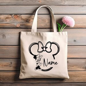May include: A white canvas tote bag with black handles. The bag features a black and white outline of Minnie Mouse's head with a bow and the text "Name".