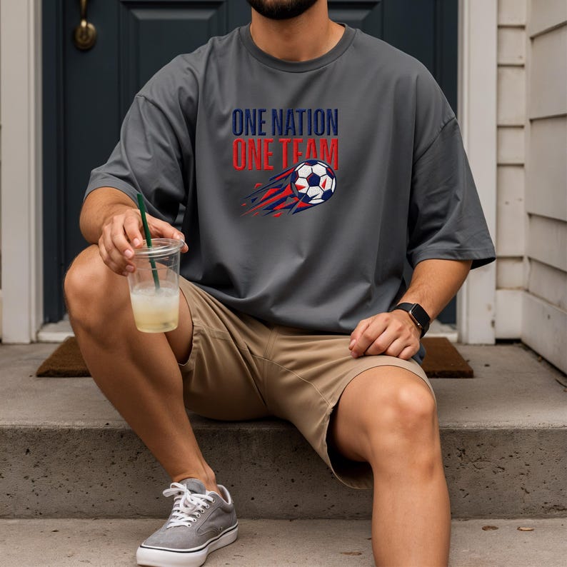 May include: Gray t-shirt with the text "ONE NATION ONE TEAM" above a soccer ball graphic with red and blue streaks. The person is wearing khaki shorts and gray sneakers.